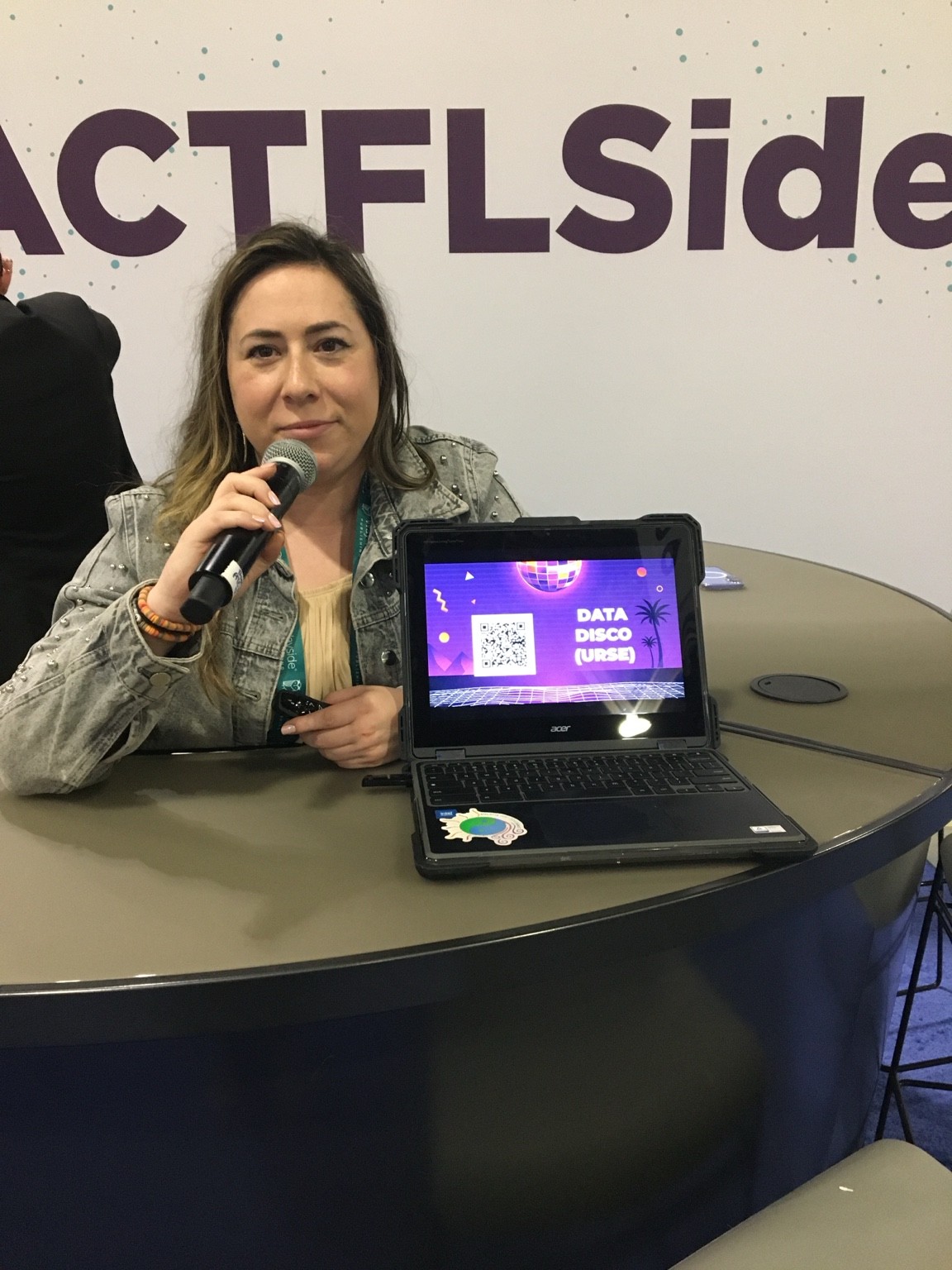 Traci Dougherty presenting with a microphone beside a conference resource display